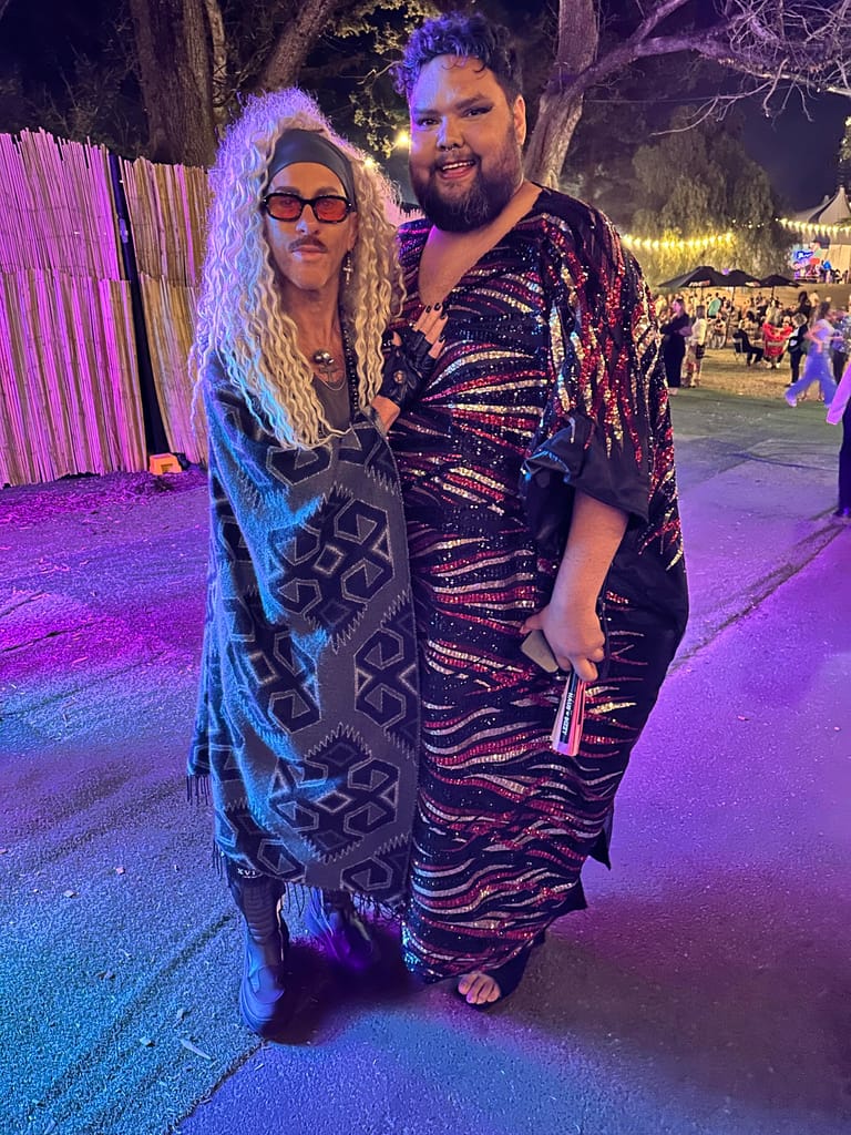 Two performers in sequinned outfits pose together under purple stage lighting at an outdoor venue.