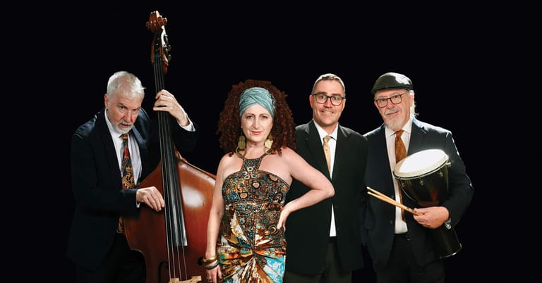 Five performers in formal attire stand against a black background, with a cellist on the left and a woman in a gold sequinned dress centre-stage.