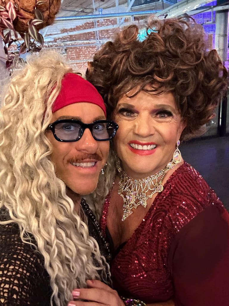 Two performers in colourful wigs and sparkly outfits smile at the camera during an Adelaide Fringe show.