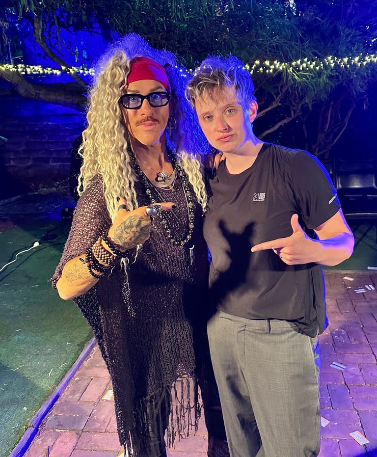 Two performers pose together under blue stage lighting, one in a sequined black outfit with long blonde hair and sunglasses, the other in a black t-shirt.