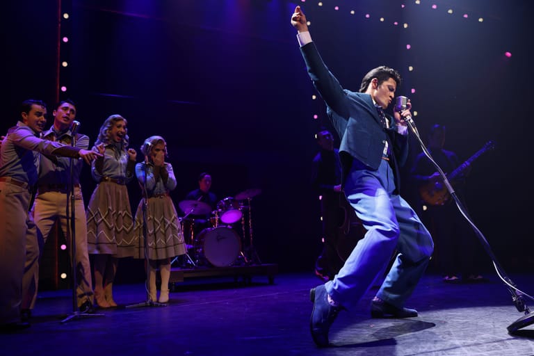 Male performer in dark suit singing with fist raised while female ensemble in gold dresses watches from stage left under blue lighting.