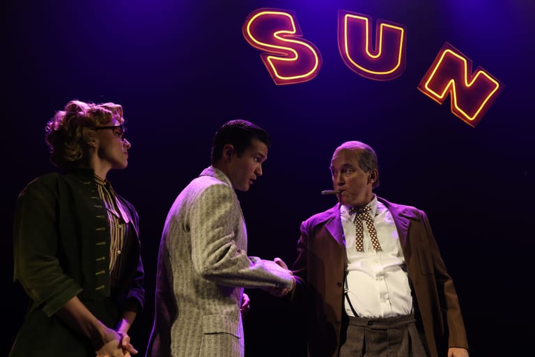 Three performers on stage in period costumes beneath a neon "SUN" sign during a theatrical production.