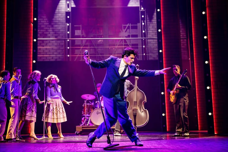 Performer in blue suit strikes a dramatic pose centre stage whilst ensemble cast stands behind under purple stage lighting.