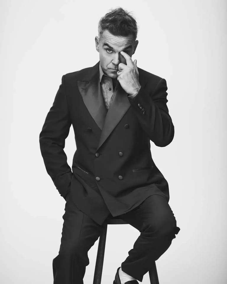 Man in black double-breasted suit seated with hand to face against white background.