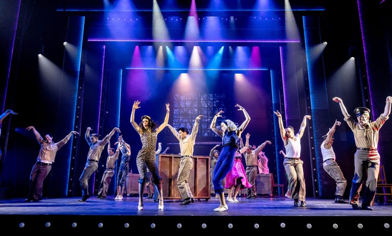 Large ensemble cast in period costumes celebrating on stage beneath blue and purple theatrical lighting.