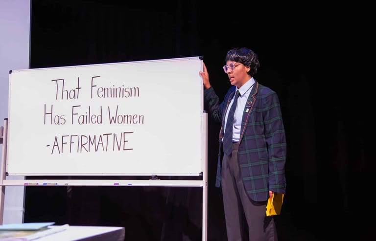 Actor in checked blazer holds protest sign reading "That Feminism Has Failed Women -AFFIRMATIVE" on darkened stage.