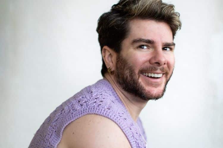 Man with dark hair and beard smiling at camera, wearing a purple knit sweater against a white background.