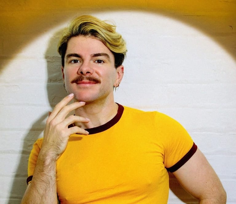 Man with blonde hair and moustache wearing yellow t-shirt, hand to chin in thoughtful pose against white wall.