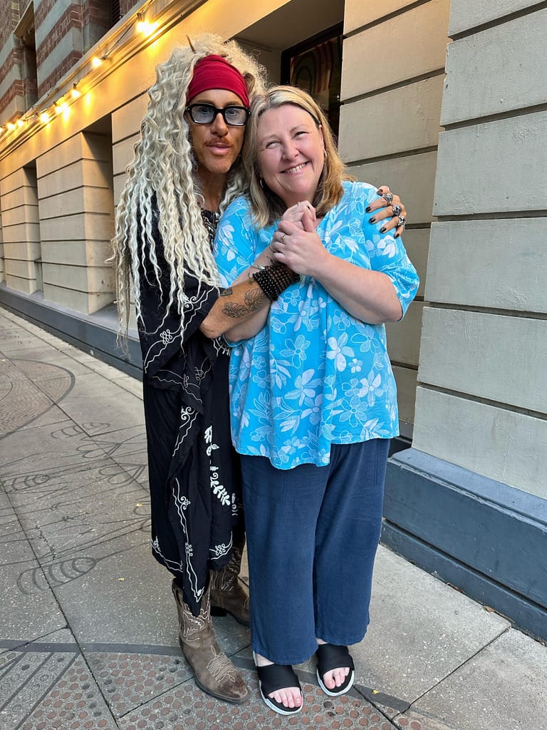 Two performers pose together on a street, one in a red cap with white dreadlocks and black outfit, the other in a blue tie-dye shirt.