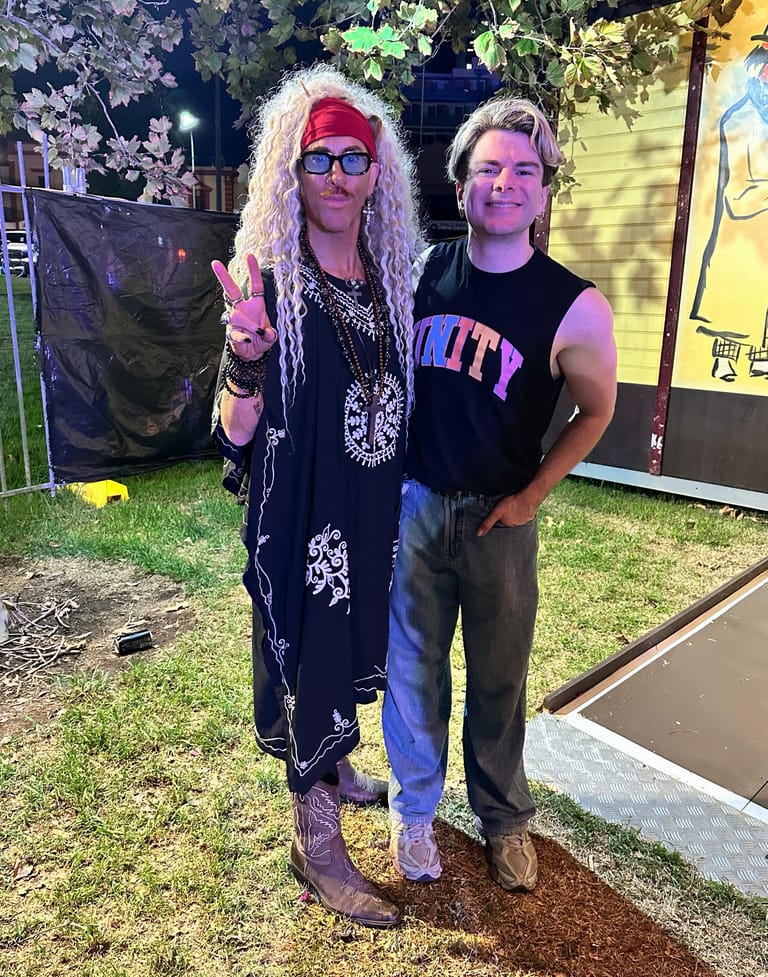 Two performers in bold stage costumes pose outdoors at night, one wearing a red cap and patterned black jacket, the other in a sleeveless black shirt.