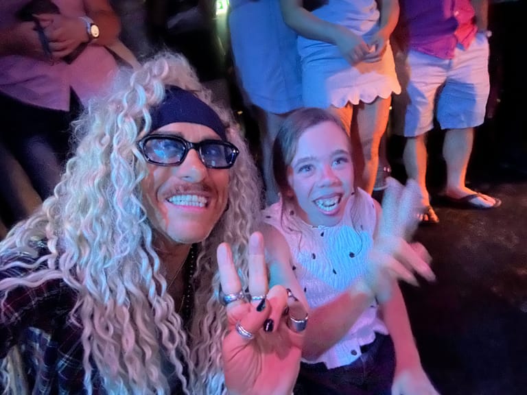 Two performers in costume and sunglasses smile at the camera during an evening event, surrounded by audience members in colourful stage lighting.