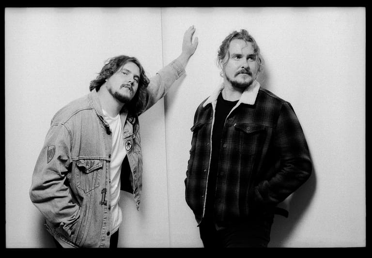 Two men stand against a white wall in black and white portrait, one gesturing upward with his arm raised.