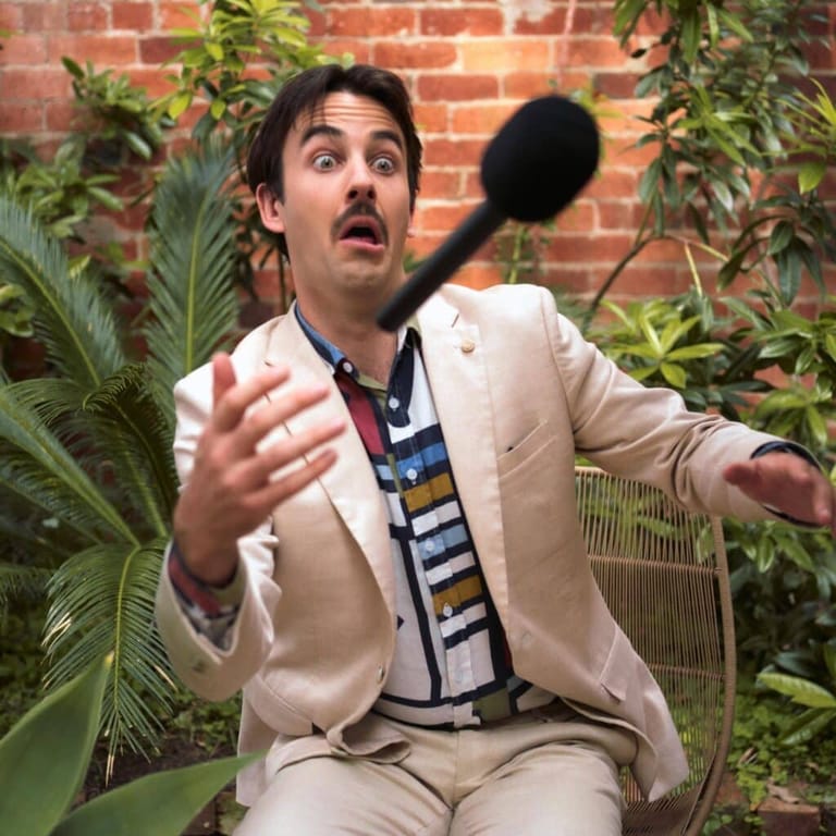Man in beige suit gestures expressively whilst seated against brick wall with climbing plants.