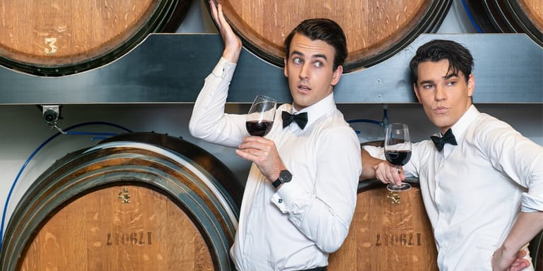 Two men in formal white shirts and bow ties hold wine glasses in front of wooden barrels.