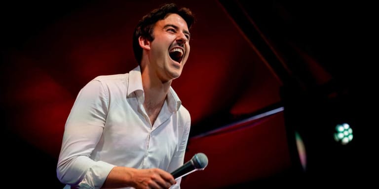 Man in light blue shirt speaking animatedly into microphone against red stage lighting.