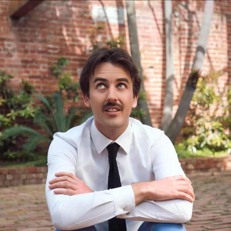 Man in white shirt and black tie with arms crossed against a brick wall backdrop.