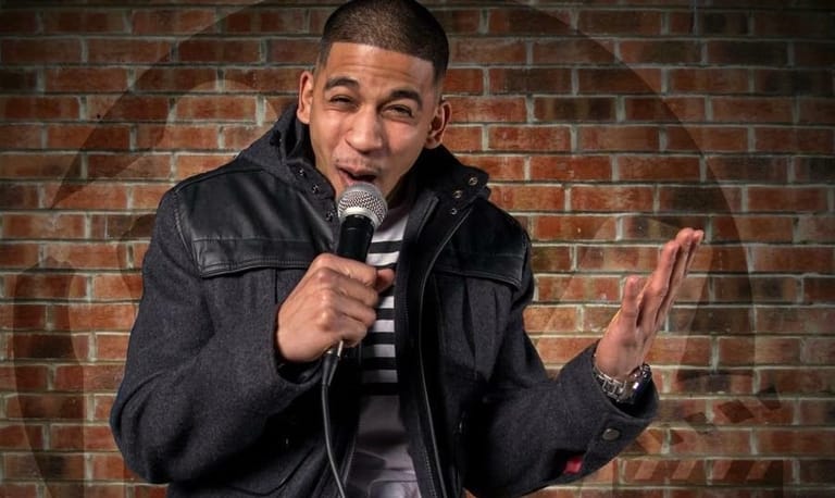Comedian holding microphone with expressive hand gesture against a brick wall backdrop.
