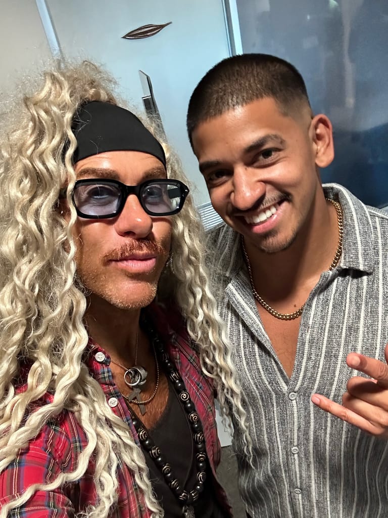 Two people pose together, one wearing long blonde wavy hair, dark sunglasses and a headband, the other in a striped shirt, both smiling at the camera.