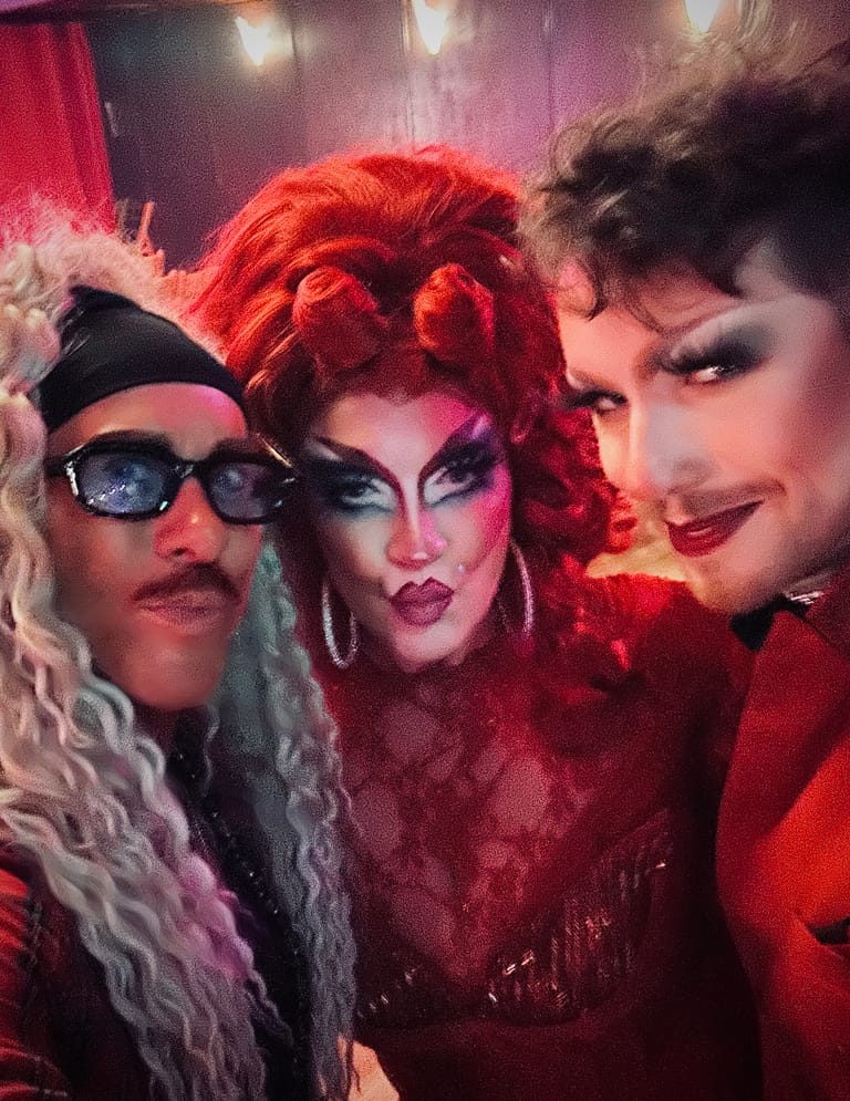 Three performers in bold makeup and wigs pose under red stage lighting during a drag production.