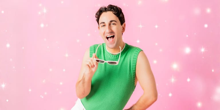 Man in a bright green sleeveless shirt holds a glowing star prop against a pink backdrop with twinkling lights.