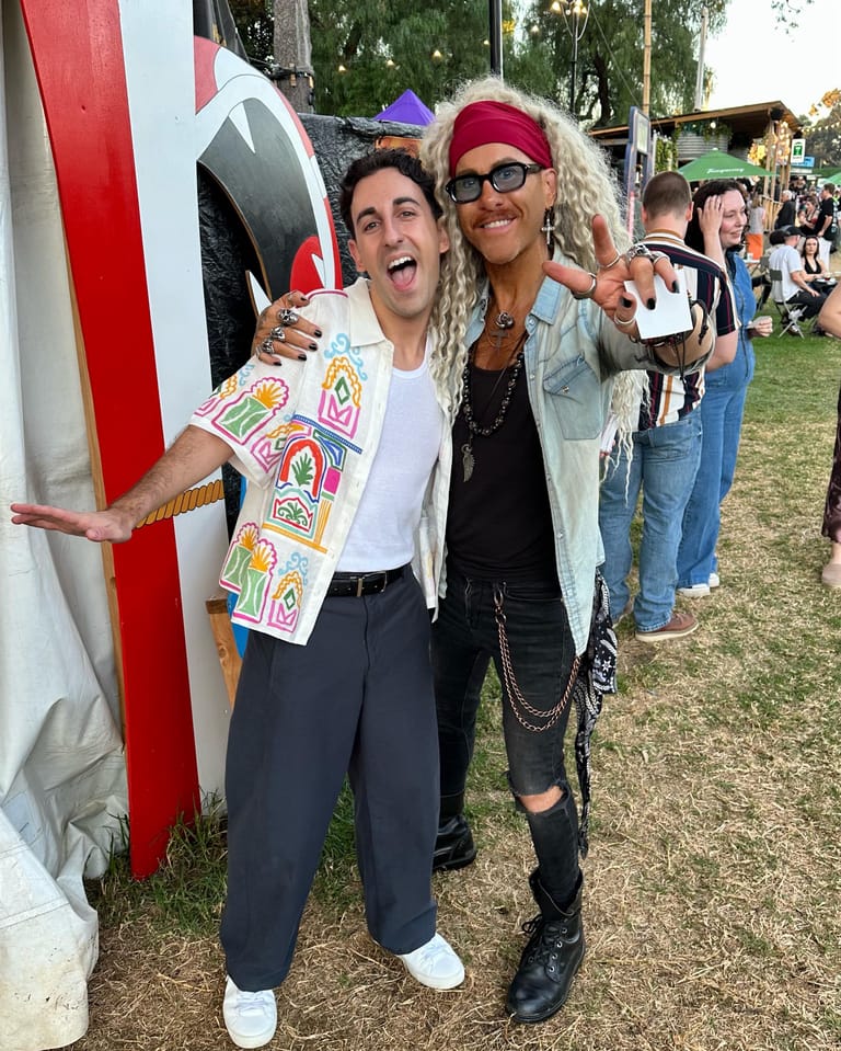 Two performers in colourful costumes pose together outdoors at a festival, smiling at the camera.
