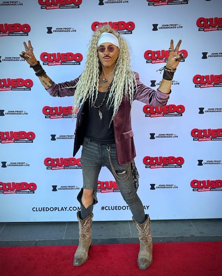 Person in long blonde dreadlocks, sunglasses and burgundy jacket poses with arms raised on Cluedo The Play red carpet.