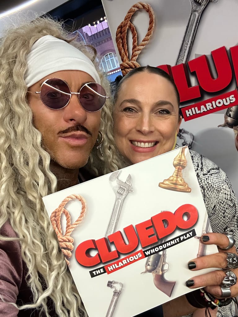 Two performers in character costumes pose together holding a Cluedo play programme, surrounded by oversized props including a wrench and rope.