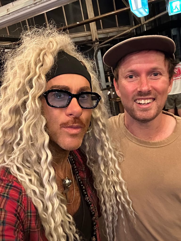 Two performers pose together, one wearing long blonde wavy hair, sunglasses and plaid shirt, the other in a beige shirt and cap.