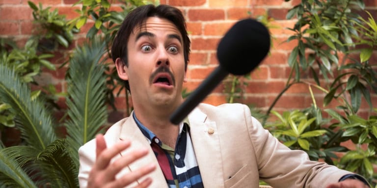 Performer in a cream blazer and blue shirt gestures expressively against a brick wall lined with potted plants.