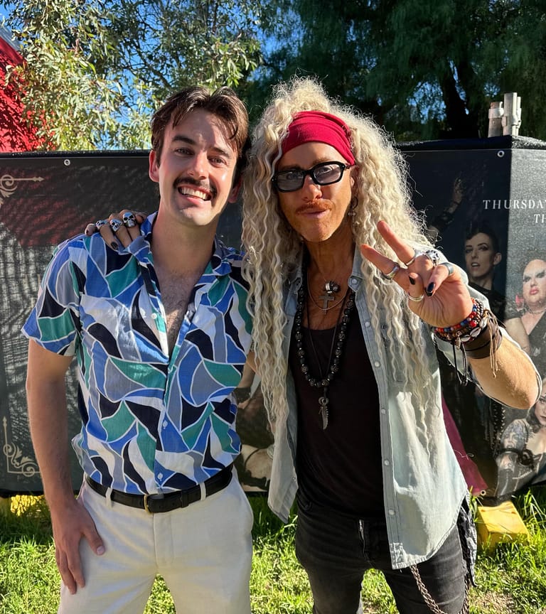 Two performers pose together outdoors in summer sunlight, one wearing a patterned blue shirt and the other a red headband with long blonde hair.