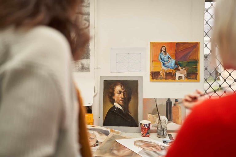 Two people view artworks displayed on a studio wall, including a portrait and colourful figurative paintings.