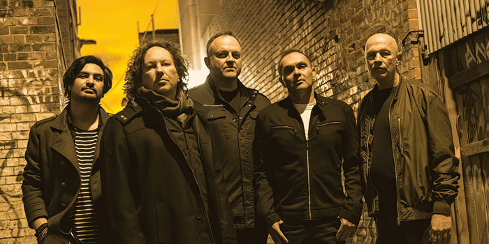 Five band members in dark jackets stand together against a brick wall lit by warm yellow light.
