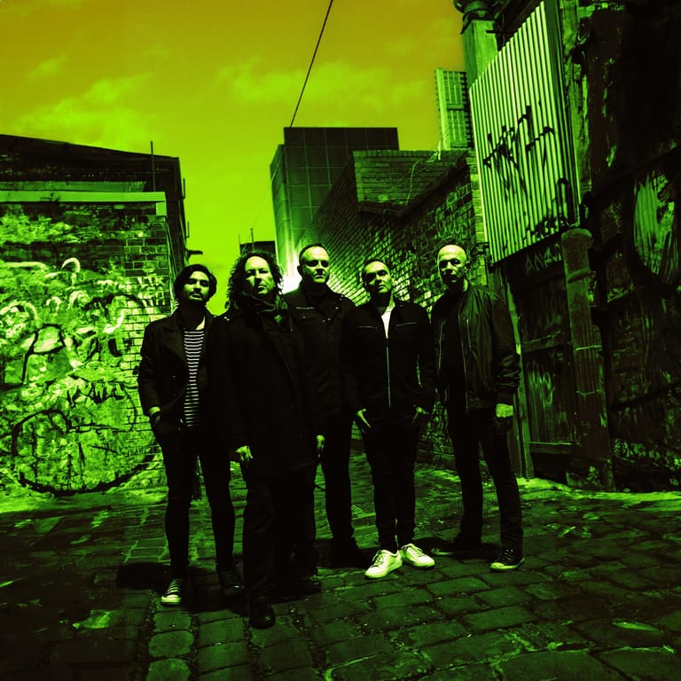 Seven band members stand in formation against neon green industrial lighting and corrugated metal backdrop.