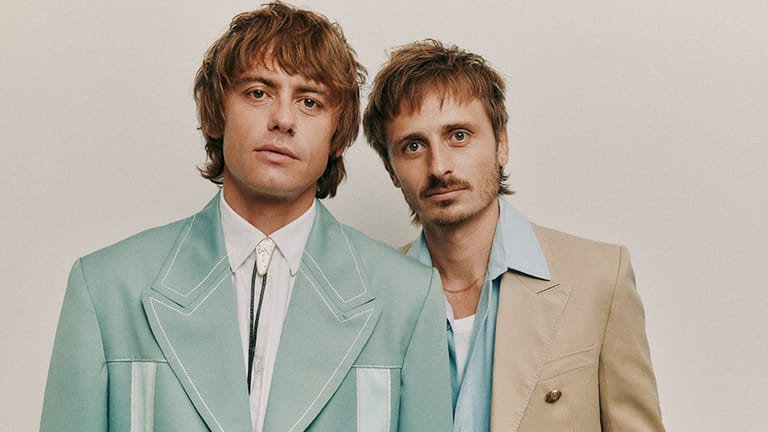 Two men in vintage-style blazers stand shoulder-to-shoulder against a neutral background, gazing directly at the camera.