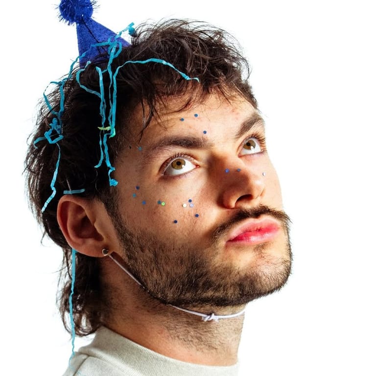 Person wearing a blue party hat with decorative strings, looking upward against a white background.