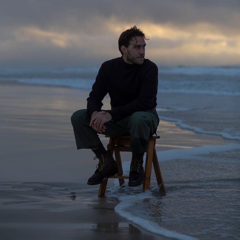 Man in dark clothing seated on a wooden chair in shallow ocean water at dusk.