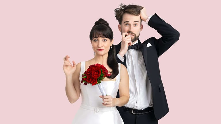 Couple in formal wedding attire holding a red rose bouquet against a pink background.