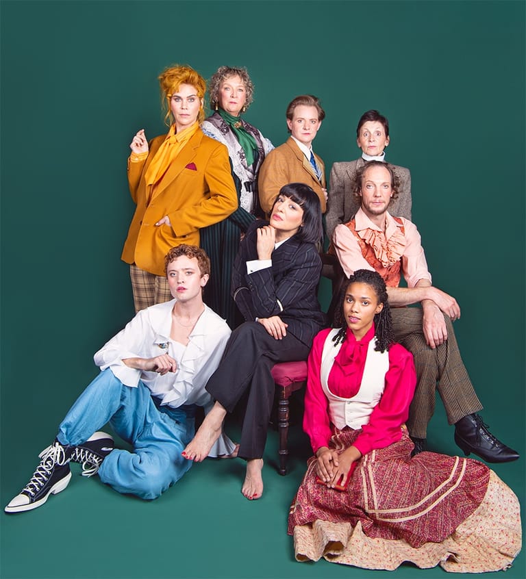 Seven people in vintage clothing posed together against a teal backdrop for a group portrait.