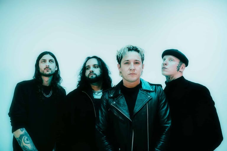 Four band members in black leather jackets stand against a pale blue backdrop, looking toward the camera.