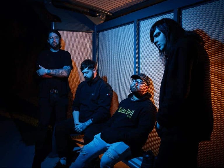 Four musicians in dark clothing stand and sit in a blue-lit room during a tour stop.