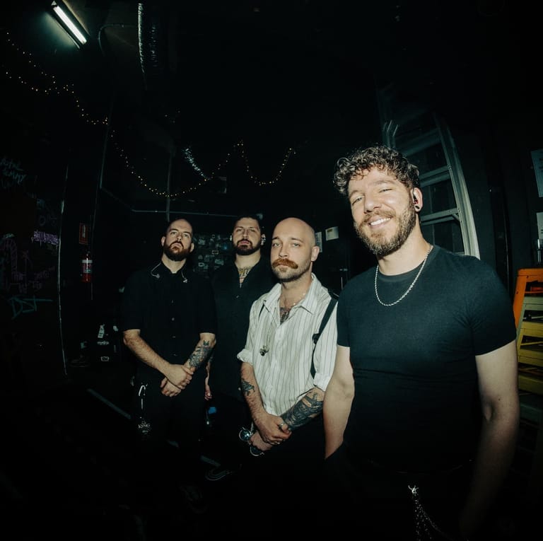 Four bearded men stand together backstage in dim lighting during a tour stop.