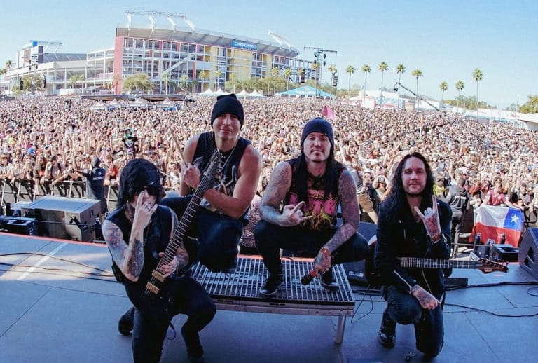 Rock band performing on stage before a massive crowd at a beachside venue during daytime.