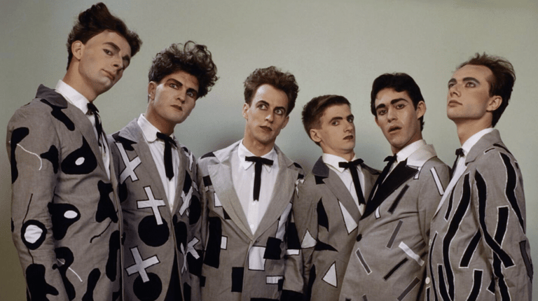 Six male performers in coordinated patterned and solid suits stand together against a neutral backdrop.
