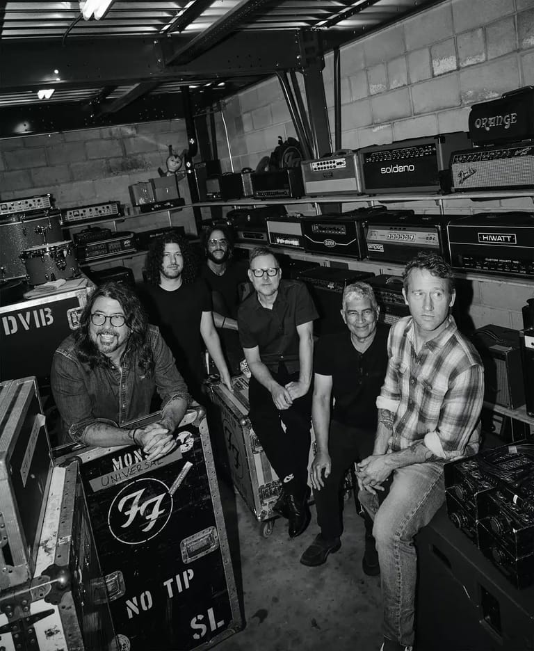 Five band members pose in a black-and-white studio surrounded by amplifiers and musical equipment.