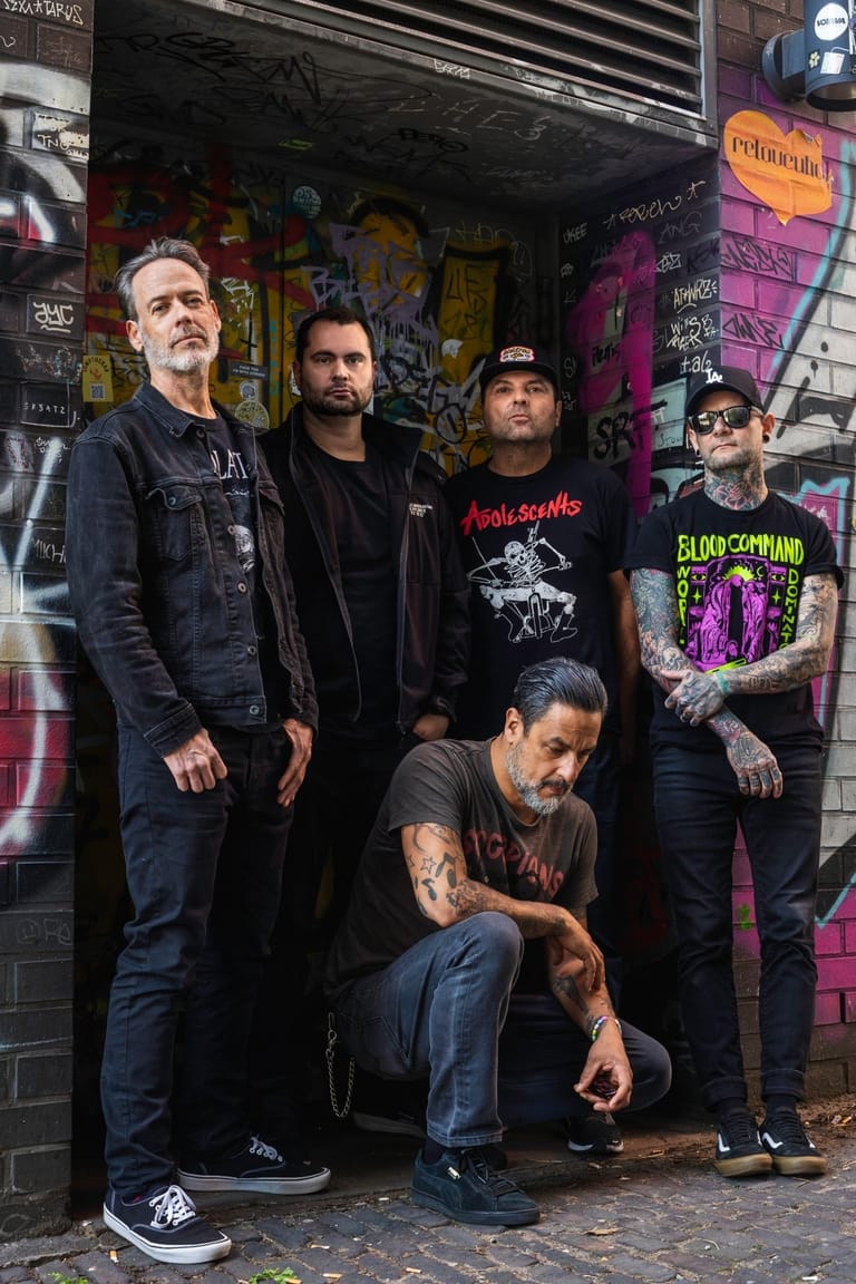 Five band members posed against a colourful graffitied brick wall in an urban laneway setting.