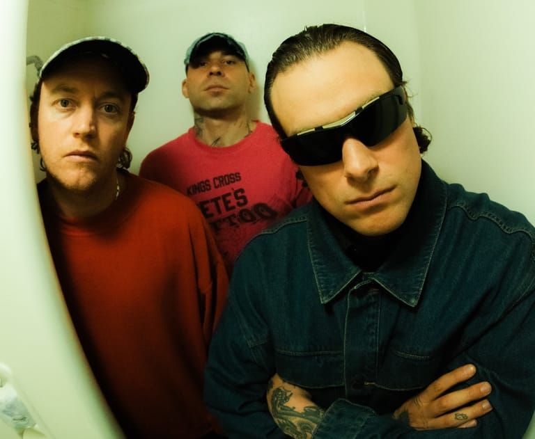 Three band members pose against a pale wall, the front figure wearing dark sunglasses and a denim shirt.