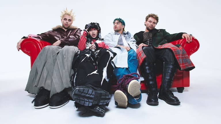 Five band members posed together on a red velvet sofa against a white background.