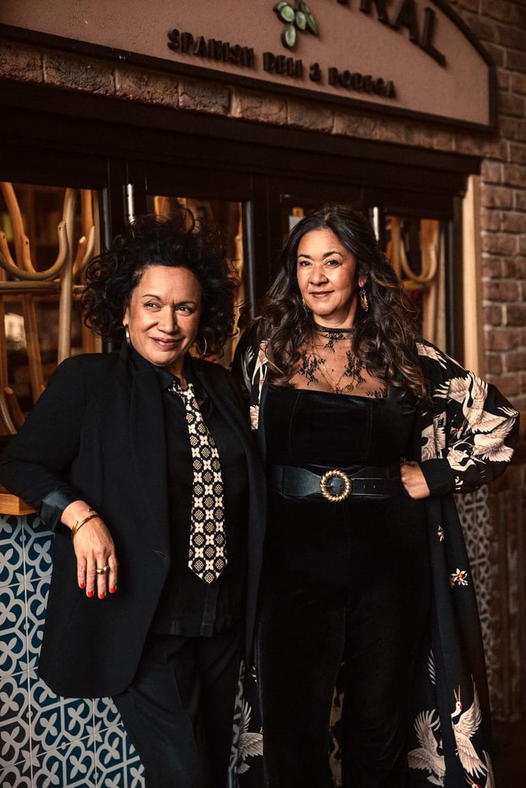 Two women in elegant black attire pose outside a theatre venue with gold-trimmed entrance doors.