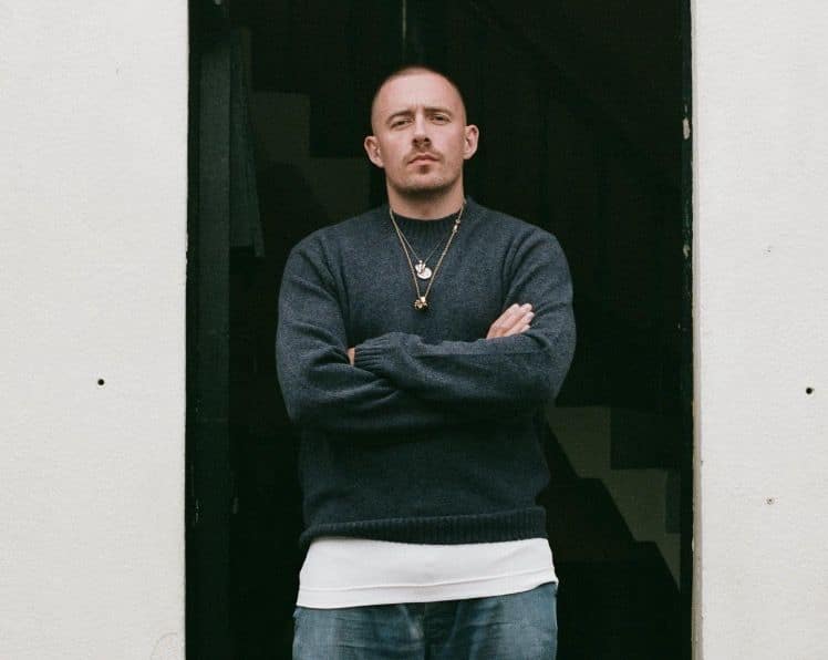 Man in dark sweatshirt and chains standing with arms crossed against a dark doorway.