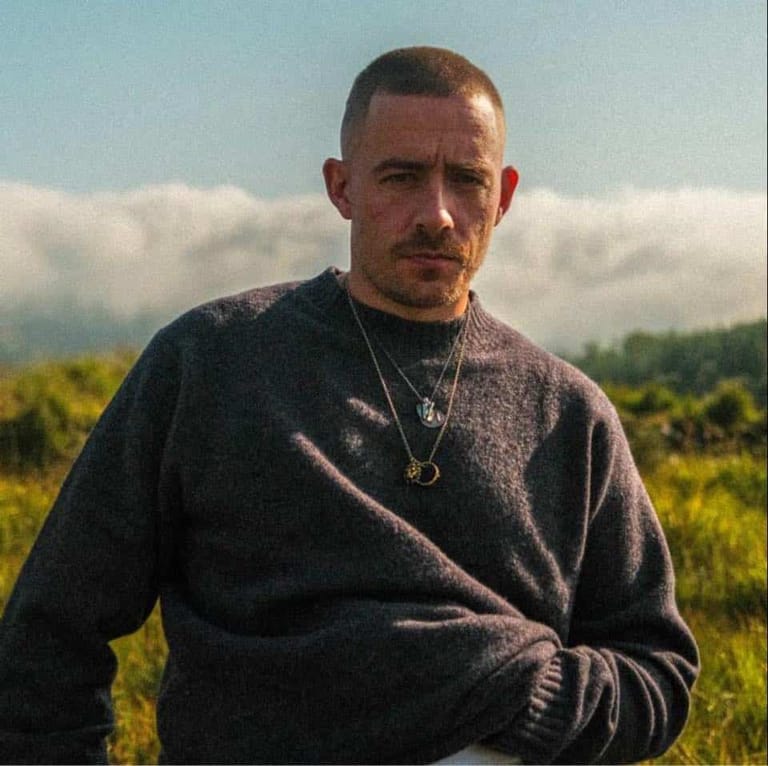 Man in dark sweatshirt standing in sprawling green field beneath cloudy sky.